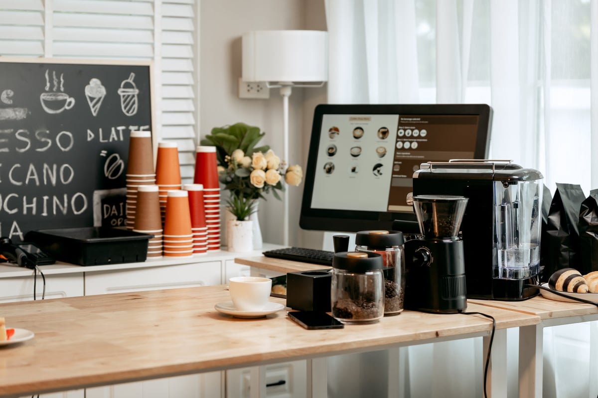 A digital coffee menu on screen at a modern café counter, surrounded by takeaway cups and brewing equipment