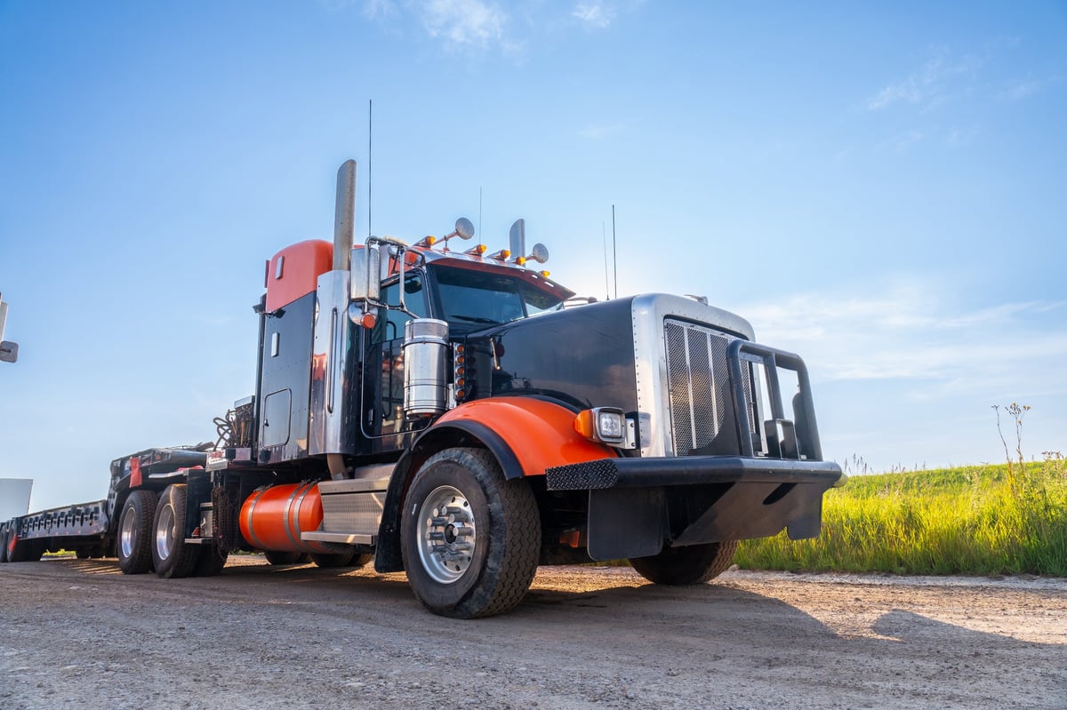 A black and orange semi-truck parked on a dirt road beside a green field under a clear blue sky, the kind of commercial vehicle CBB Group arranges truck and equipment financing for